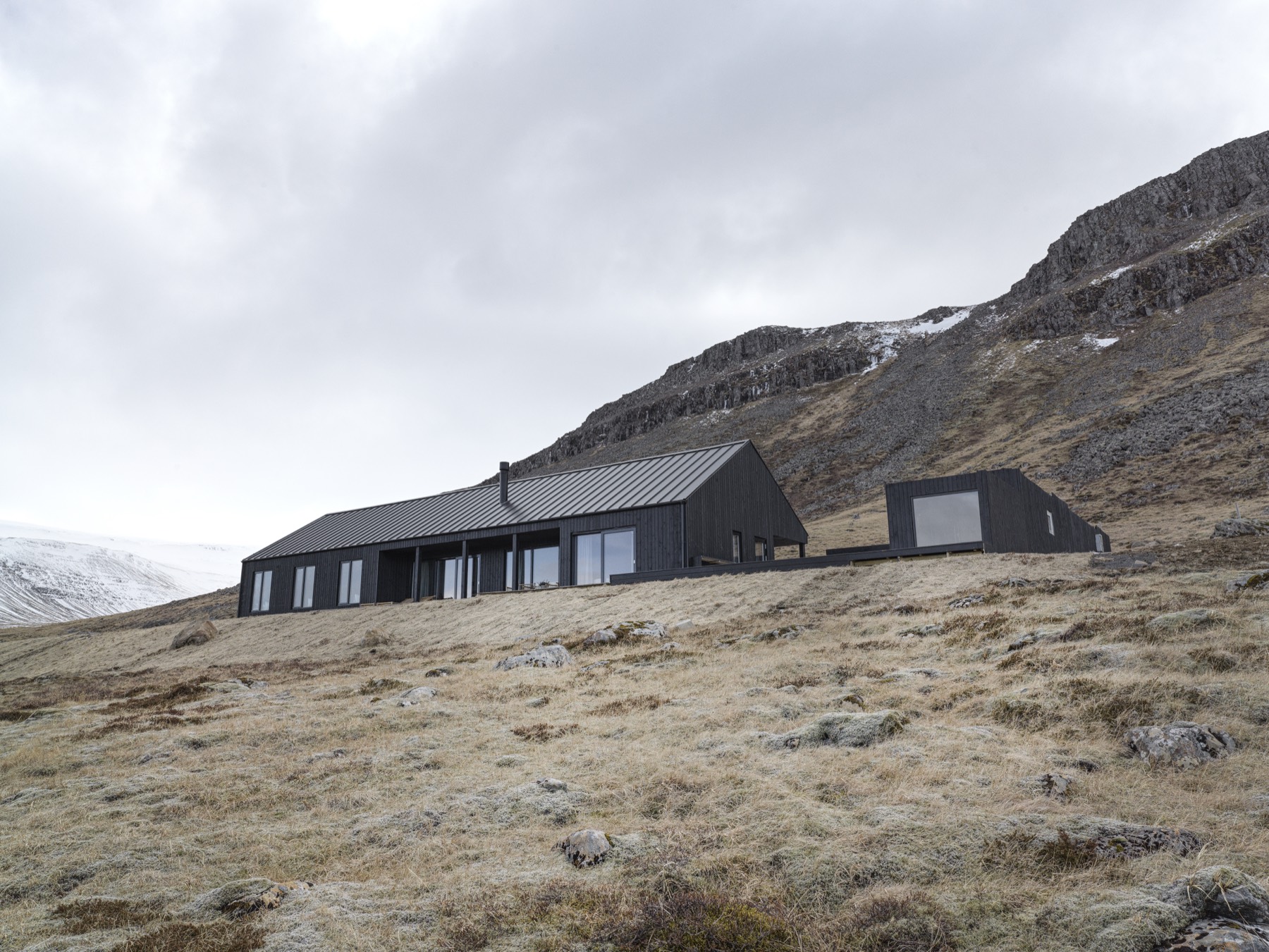 The Lake House at Meðalfellsvatn — black timber-clad volumes set into a hillside below a snow-dusted ridge.